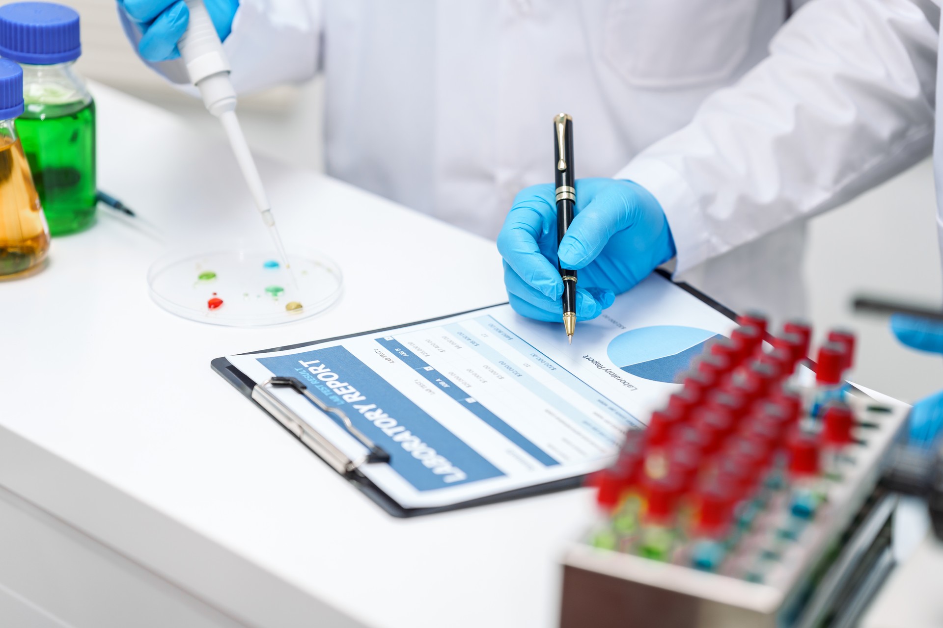 Two Asian scientists in lab coats sit at a table in a laboratory, examining samples through a microscope. The room is equipped with scientific tools, liquids, and chemistry materials.